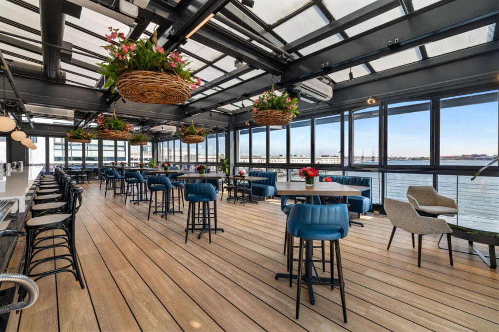 A stylish indoor restaurant with large windows offering a view of the water. The seating includes blue cushioned chairs at tall round tables and bar stools. The decor features hanging baskets with flowers and modern lighting, creating a bright and inviting atmosphere.