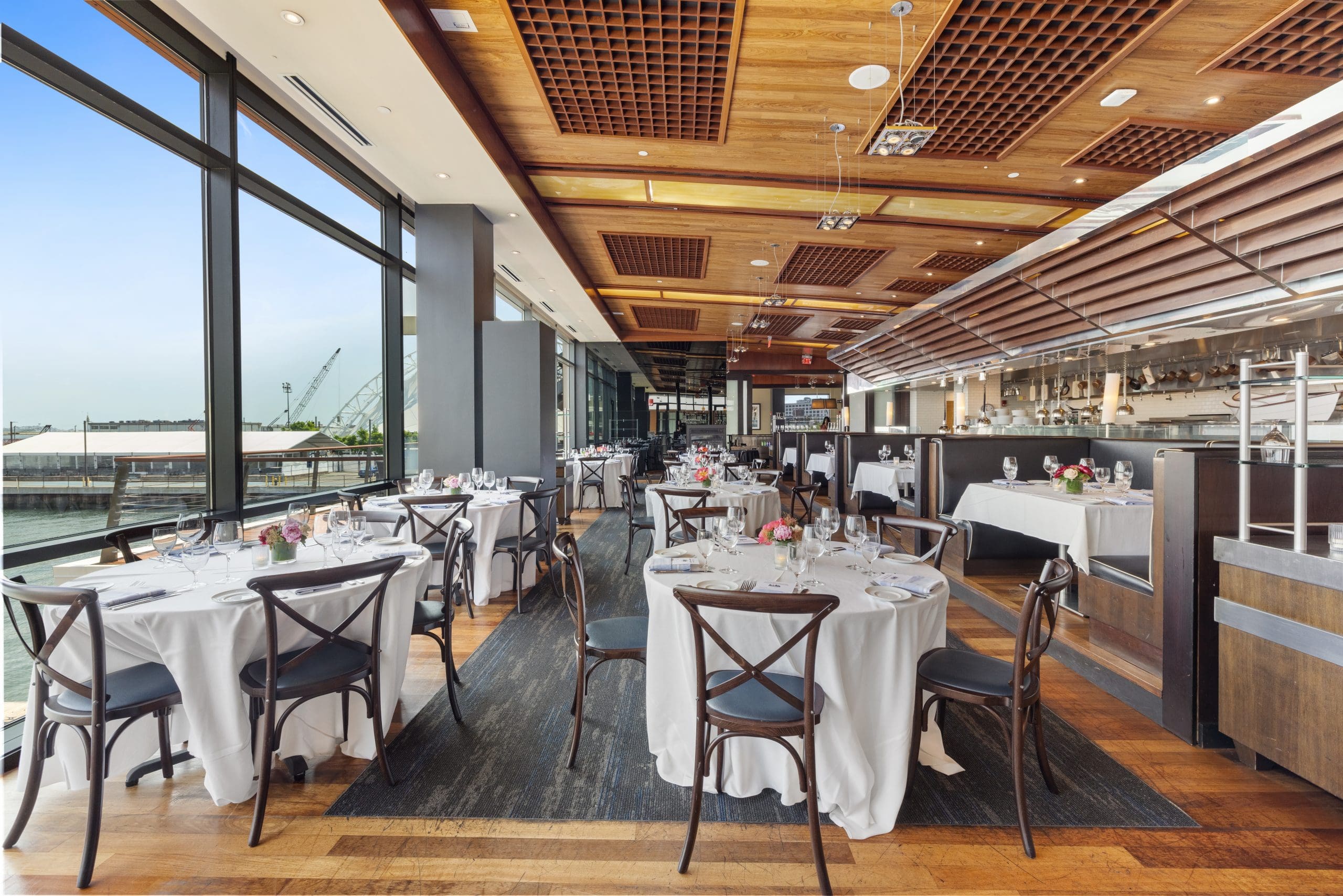 A modern restaurant with large windows offering a waterfront view. Tables are set with white tablecloths, silverware, and centerpieces featuring pink flowers. The ceiling has wooden accents with recessed lighting, and the interior is spacious and well-lit.