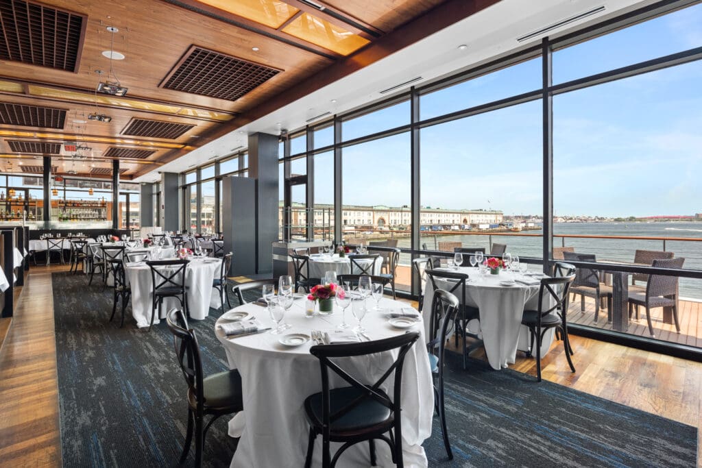 A spacious dining room with round tables covered in white tablecloths, set with polished cutlery and glassware, and small red flower centerpieces. Floor-to-ceiling windows offer a scenic waterfront view. Black wooden chairs surround the tables.