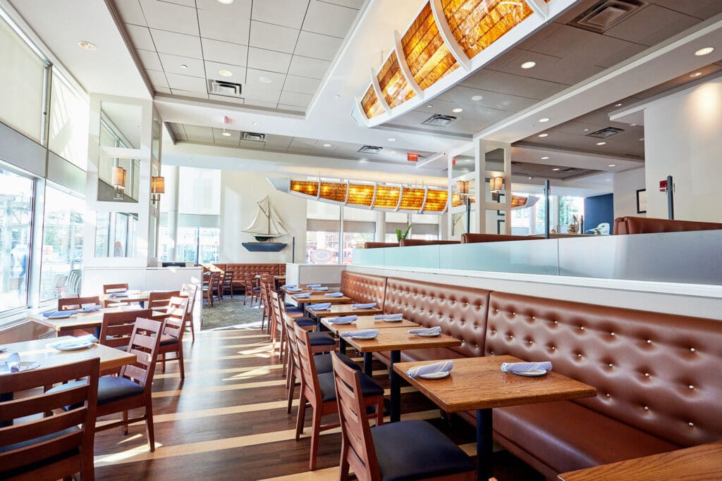 A bright, modern restaurant features wooden tables and chairs, with a long tufted booth along one wall. Large windows allow natural light to flood in. Warm lighting from overhead fixtures and a nautical-themed decor, including a model sailboat, complete the setting.