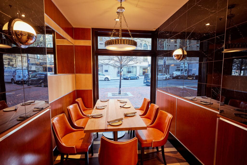 A cozy, small restaurant dining area features a wooden table set for six with elegant orange leather chairs. Large windows offer a view of the street outside, allowing natural light to fill the space. The walls are a mix of wooden panels and black marble.