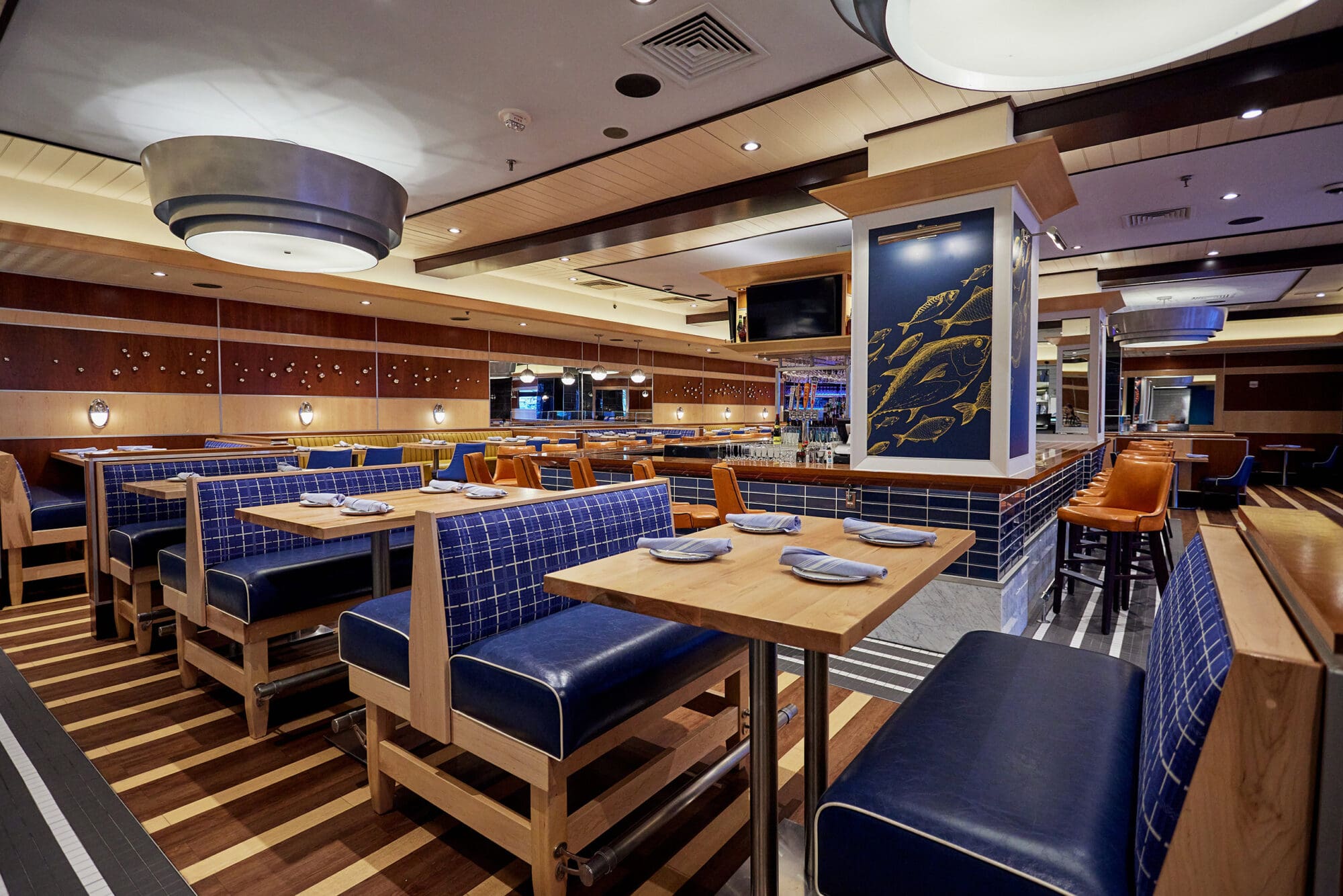 A modern, well-lit restaurant interior with blue and wooden booth seating on the left, set tables, and bar stools along the right side. The decor includes a nautical theme with fish artwork and warm wood accents throughout the space.