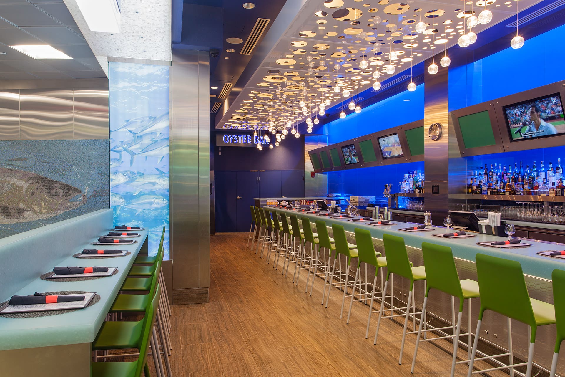 A modern seafood bar with a nautical theme. The bar has green barstools, an illuminated blue backdrop, and hanging circular decorations on the ceiling. Menu placards and black napkins on place settings adorn the countertop. TVs display sport channels.