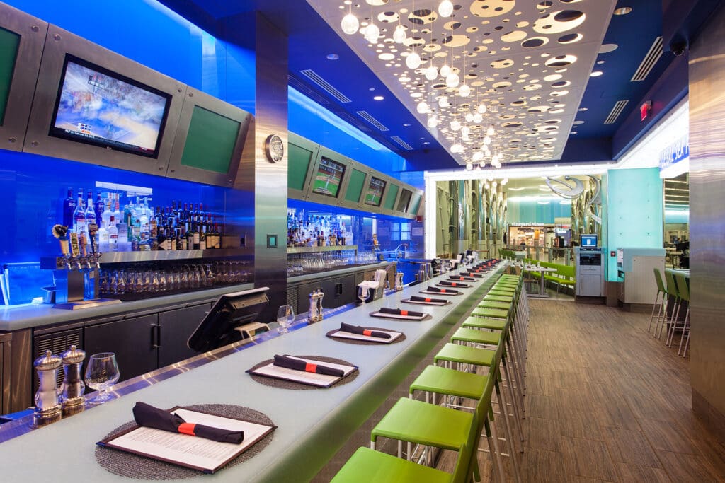 A modern bar with a long countertop lined with green bar stools. The ceiling features artistic light fixtures, and there are multiple wall-mounted TVs. The countertop has neatly arranged place settings with black napkins and utensils. The ambiance is sleek and stylish.