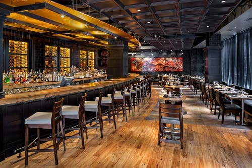 A modern restaurant interior with a long wooden bar counter on the left, topped with stools, and dining tables with chairs neatly arranged on the right. The room features warm lighting, a mix of wooden and dark tones, and a large colorful mural on the back wall.