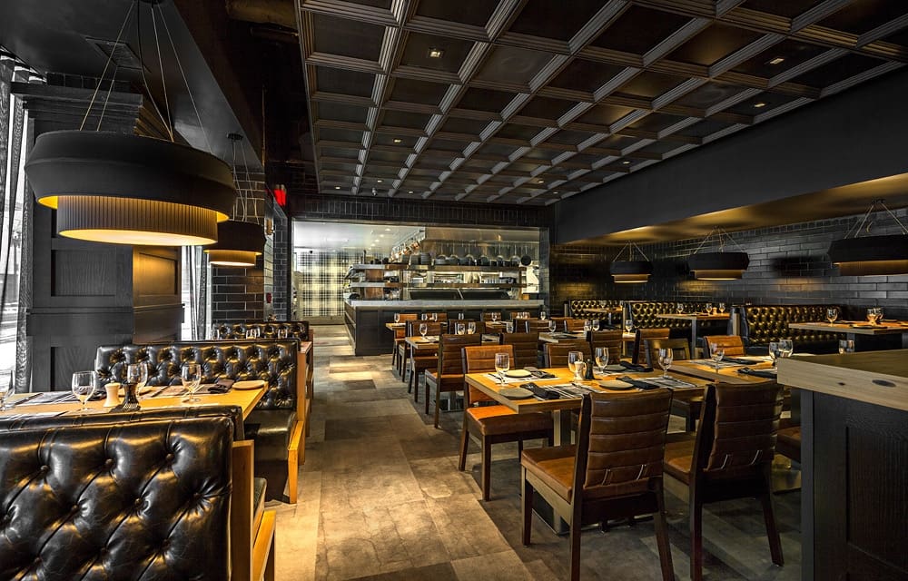 A modern, dimly lit restaurant with dark leather tufted booths and wooden chairs around neatly set tables. Large black pendant lights hang from the ceiling, and the open kitchen is visible in the background. The ambiance is elegant and cozy.