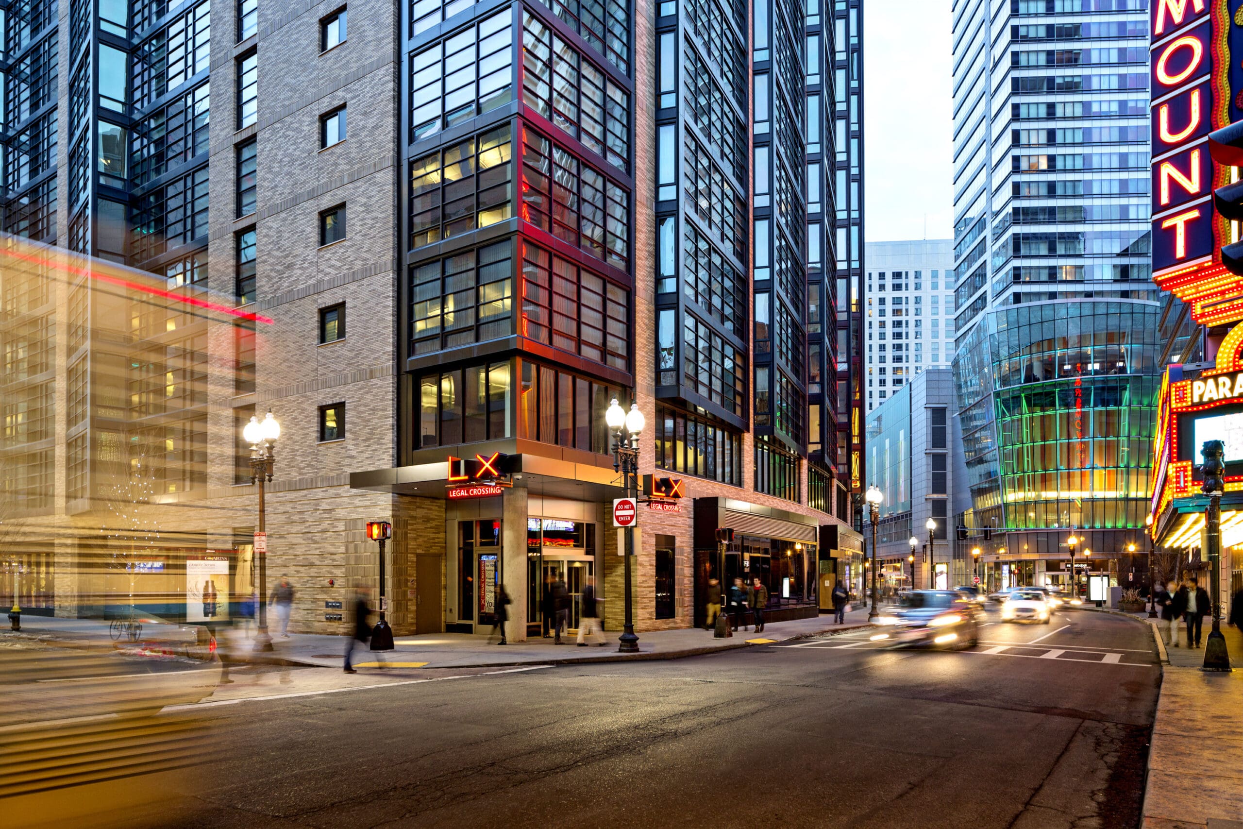 A city street at dusk featuring busy traffic and pedestrians. High-rise buildings with modern glass and brick architecture line the street. A brightly lit theater marquee on the right and motion-blur effects from passing cars and people add dynamic elements.