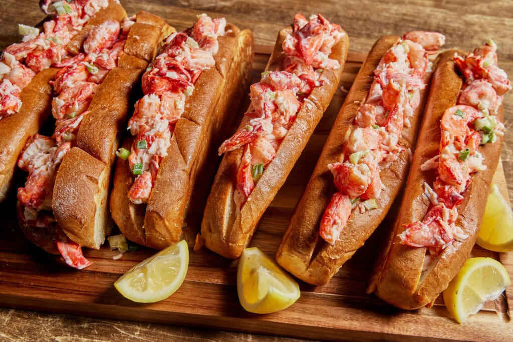 A wooden board holds five lobster rolls filled with generous chunks of lobster meat, garnished with green herbs. Lemon wedges are placed around the rolls, adding a fresh touch to the seafood presentation. The rolls rest on a rustic wooden surface.