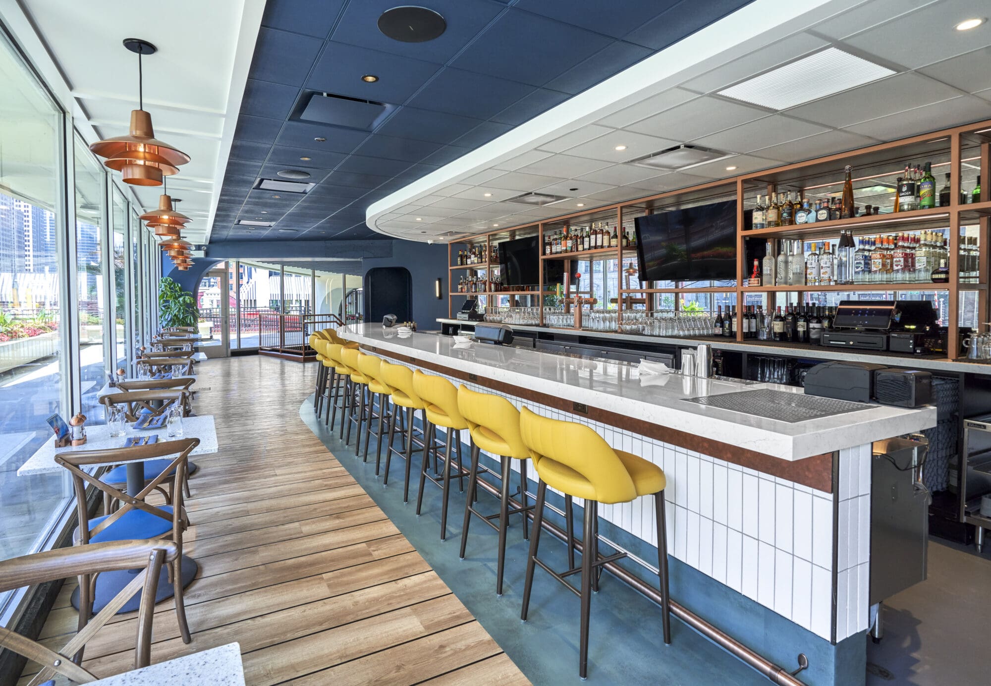 A modern bar with a long white countertop lined with bright yellow bar stools. Shelves stocked with various bottles of alcohol are visible behind the bar. The space also features small tables with chairs along a glass window, allowing natural light to fill the area.