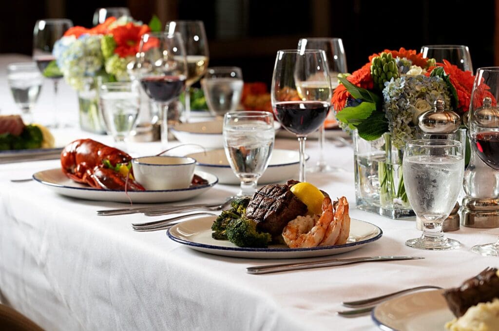 A beautifully set dining table with plates of gourmet food, including a lobster dish, a steak with shrimp, and steamed vegetables. The table is adorned with glasses of red wine and water, and decorated with vibrant flower arrangements in the center.