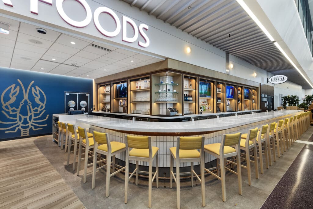 A modern seafood restaurant bar with a sleek, white countertop and rows of yellow cushioned bar stools. Behind the bar, shelves display glassware and bottles. On the left, a wall features a large lobster graphic. The ceiling has curved, ribbed panels.