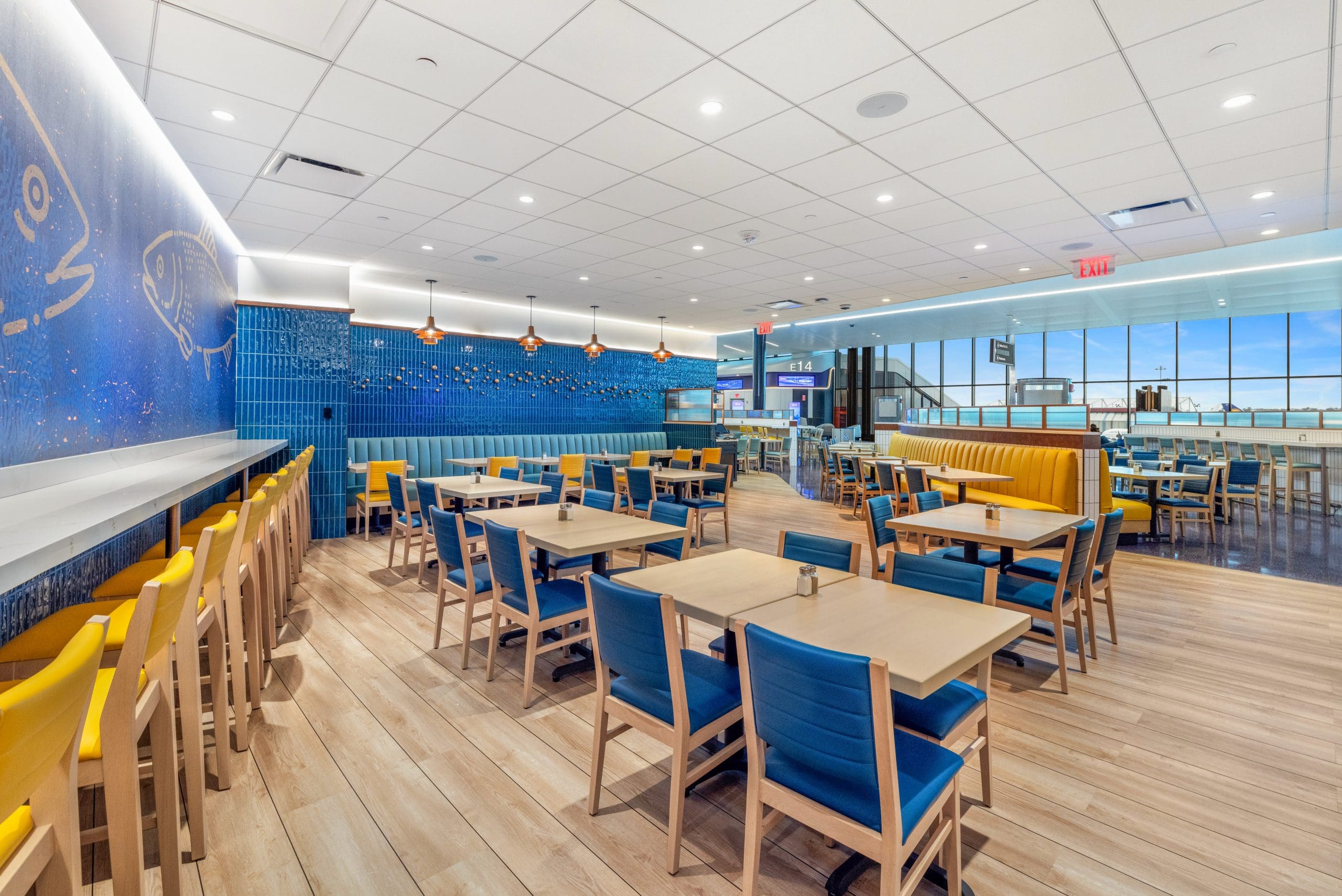 A modern restaurant interior with blue and yellow seating. The room features wooden tables, blue cushioned chairs, and yellow barstools along a counter. Fish and ocean-themed wall art decorates one side, while large windows allow natural light to fill the space.