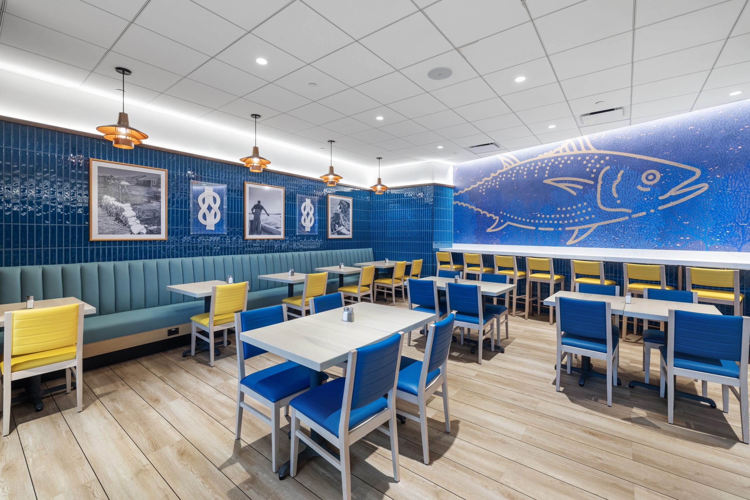 A modern restaurant interior featuring blue and yellow chairs around light wooden tables. The walls are decorated with fish-themed artwork, including a large blue fish mural. Pendant lights hang from the ceiling, and the floor has a light wood finish.