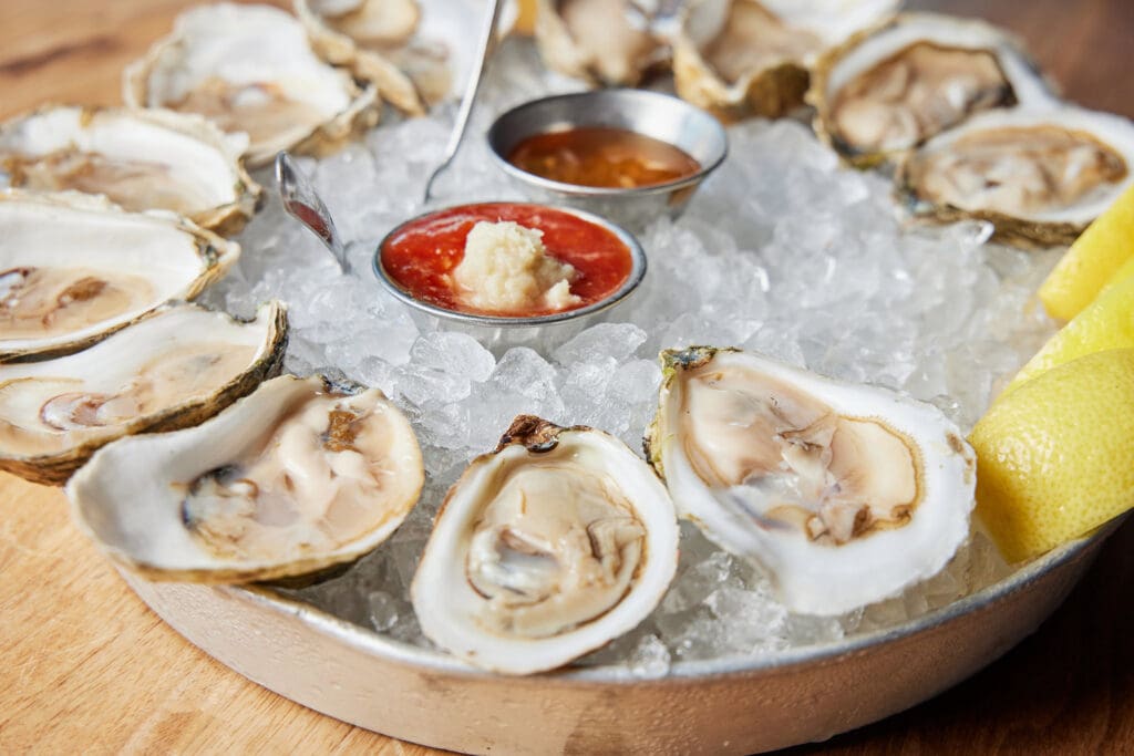 A round platter of raw oysters on a bed of ice is garnished with lemon wedges. Two small metal containers in the center hold a red cocktail sauce and a white horseradish sauce. The platter is set on a wooden surface.