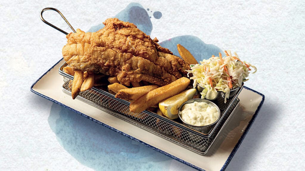 A serving of crispy fried fish laid on a bed of golden fries, accompanied by a side of creamy coleslaw. A lemon wedge and a small cup of tartar sauce complete the meal, presented in a wire basket on a square white plate with a blue border.