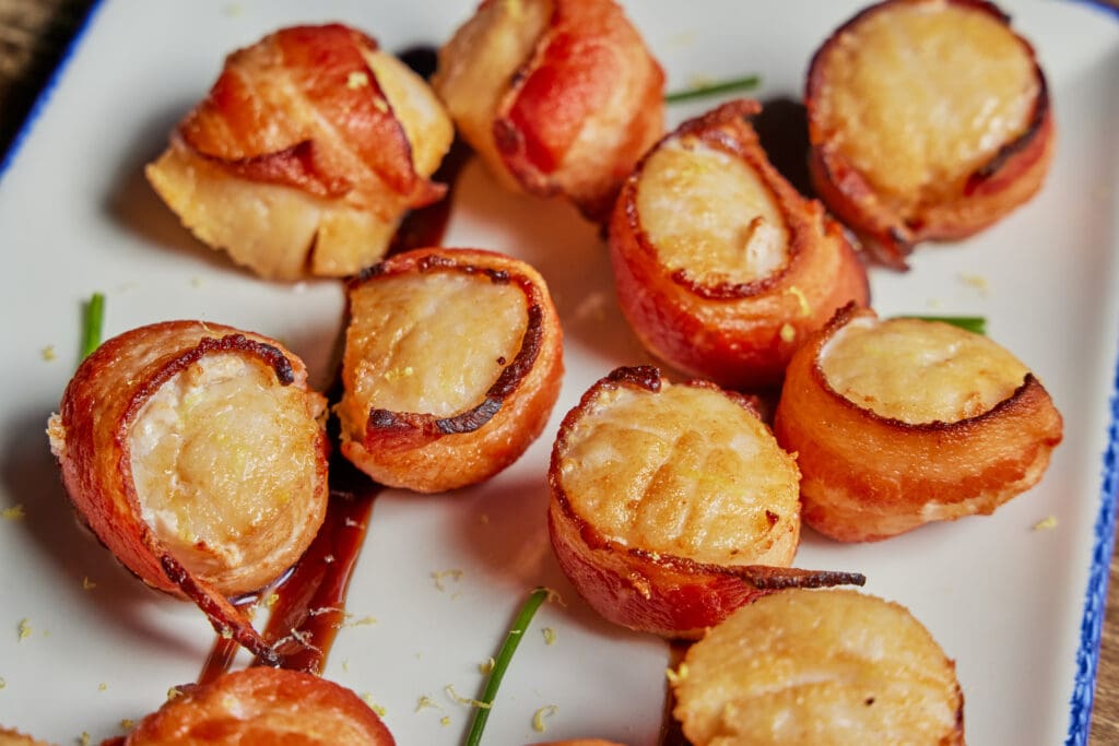 A plate of bacon-wrapped scallops garnished with chives and a sprinkle of lemon zest. The scallops are golden brown and neatly arranged on a white rectangular plate.