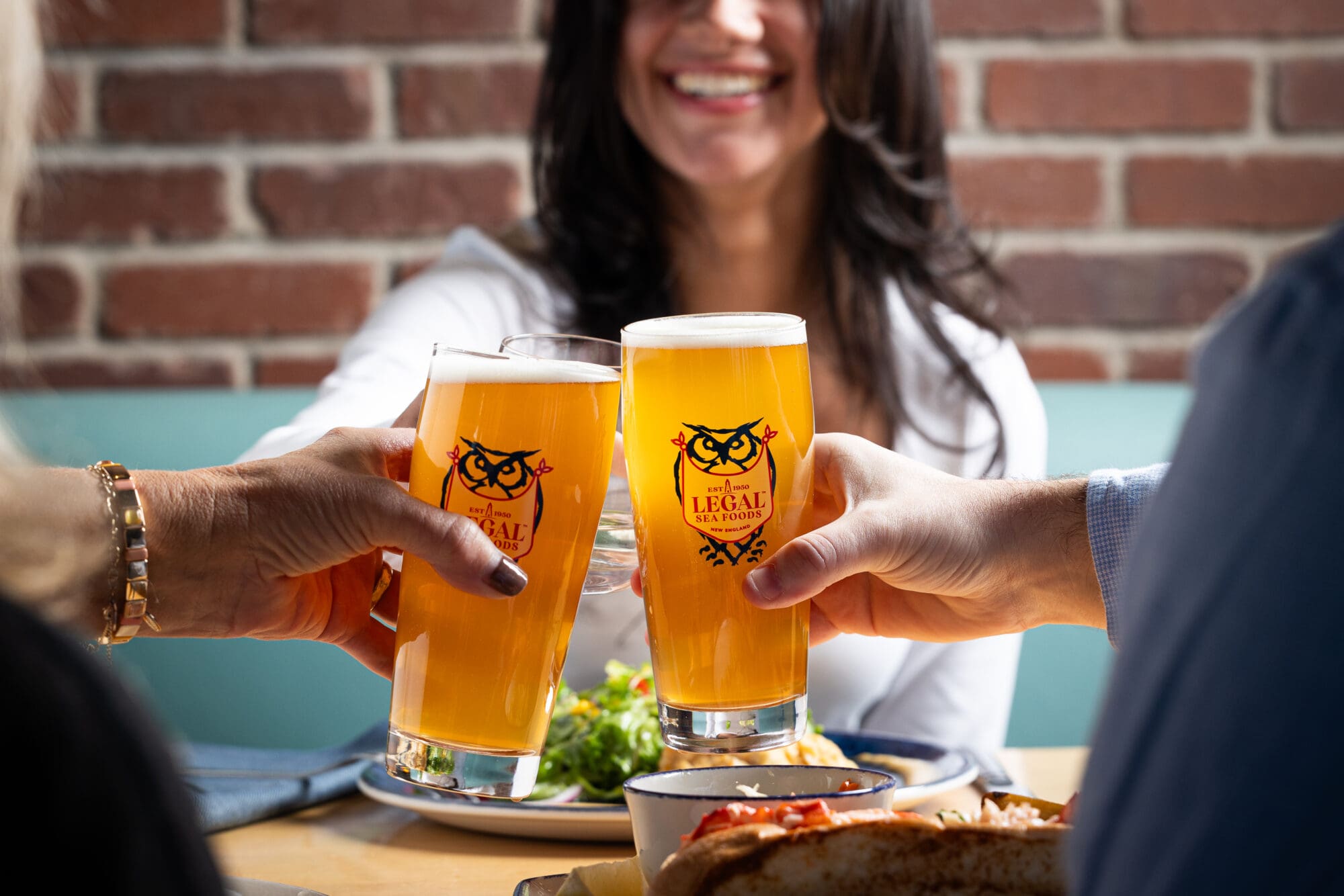 Three people clink glasses of beer in a cheerful toast at a restaurant. A woman in the background smiles broadly. The table has plates of food and a brick wall is visible behind them.