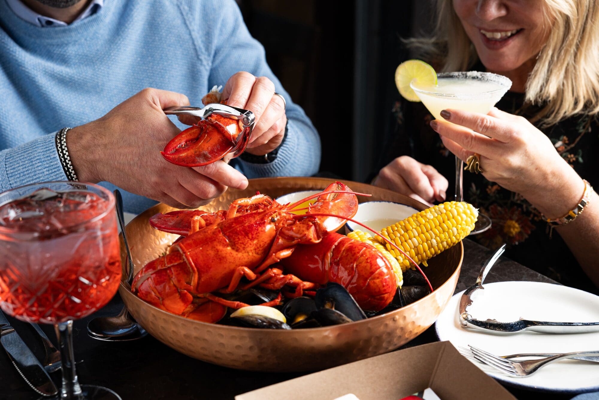 Two people enjoying a seafood meal, featuring lobsters, corn, and mussels in a copper bowl. One person is using a cracker tool, while the other holds a cocktail with a lemon garnish. Drinks are visible on the table.