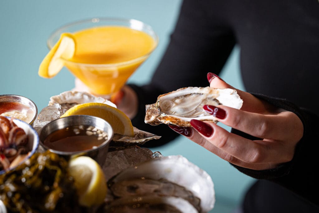 A person with red nails holds an oyster over a seafood platter, accompanied by lemon wedges and dipping sauces, with a bright orange cocktail in a martini glass nearby.