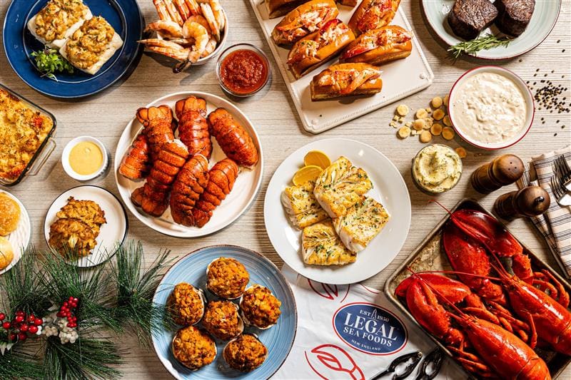 A festive spread of seafood dishes on a table, featuring lobster tails, cooked fish, crab cakes, shrimp, and clam chowder. The table is decorated with greens and pinecones, evoking a holiday theme.