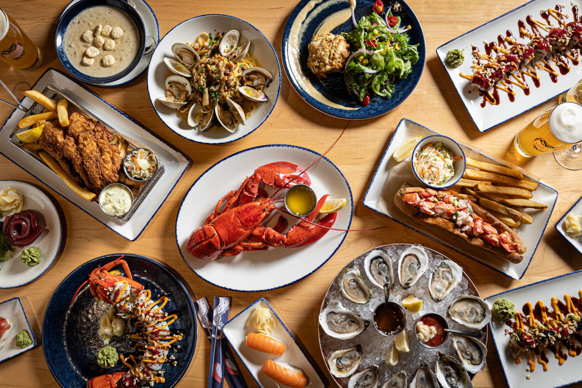 A variety of seafood dishes are spread on a wooden table, including a whole lobster, oysters, clams, sushi, and fried fish. Accompaniments like fries, salad, and sauces are also present, along with drinks.