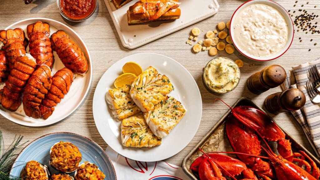 A table filled with various seafood dishes, including lobster tails, grilled fish with lemon slices, crab cakes, a whole lobster, and a bowl of creamy chowder. Garnishes and condiments are placed around the dishes.