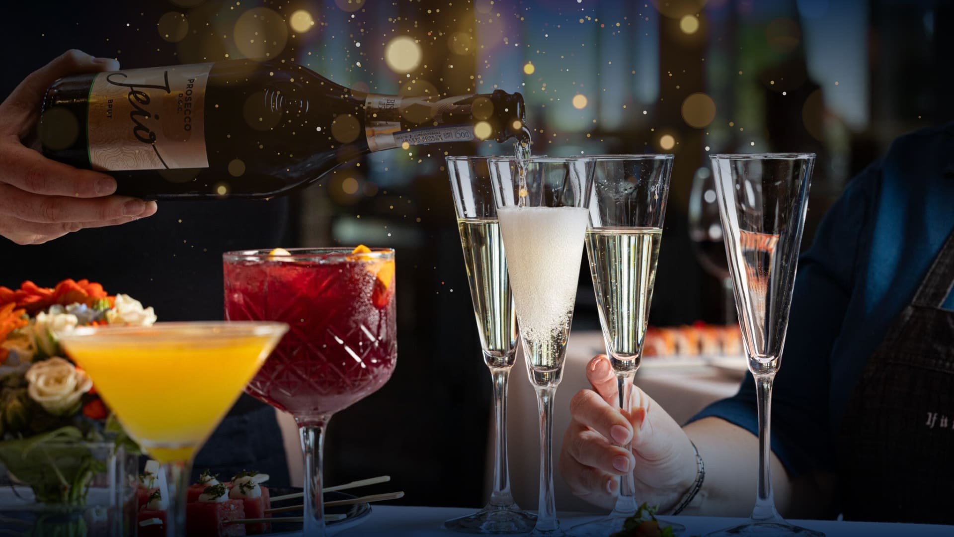 A festive setting with a hand pouring champagne into flutes. A red cocktail and a yellow-orange drink are in the foreground. Soft bokeh lights create a warm ambiance on the table, adorned with colorful flowers.