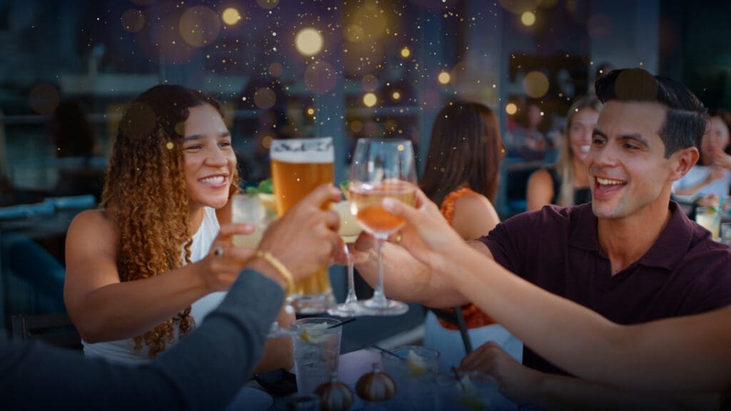 A group of people sitting outside at a restaurant cheers with various drinks, including wine and beer. They are smiling and enjoying a lively atmosphere with decorative lights in the background.