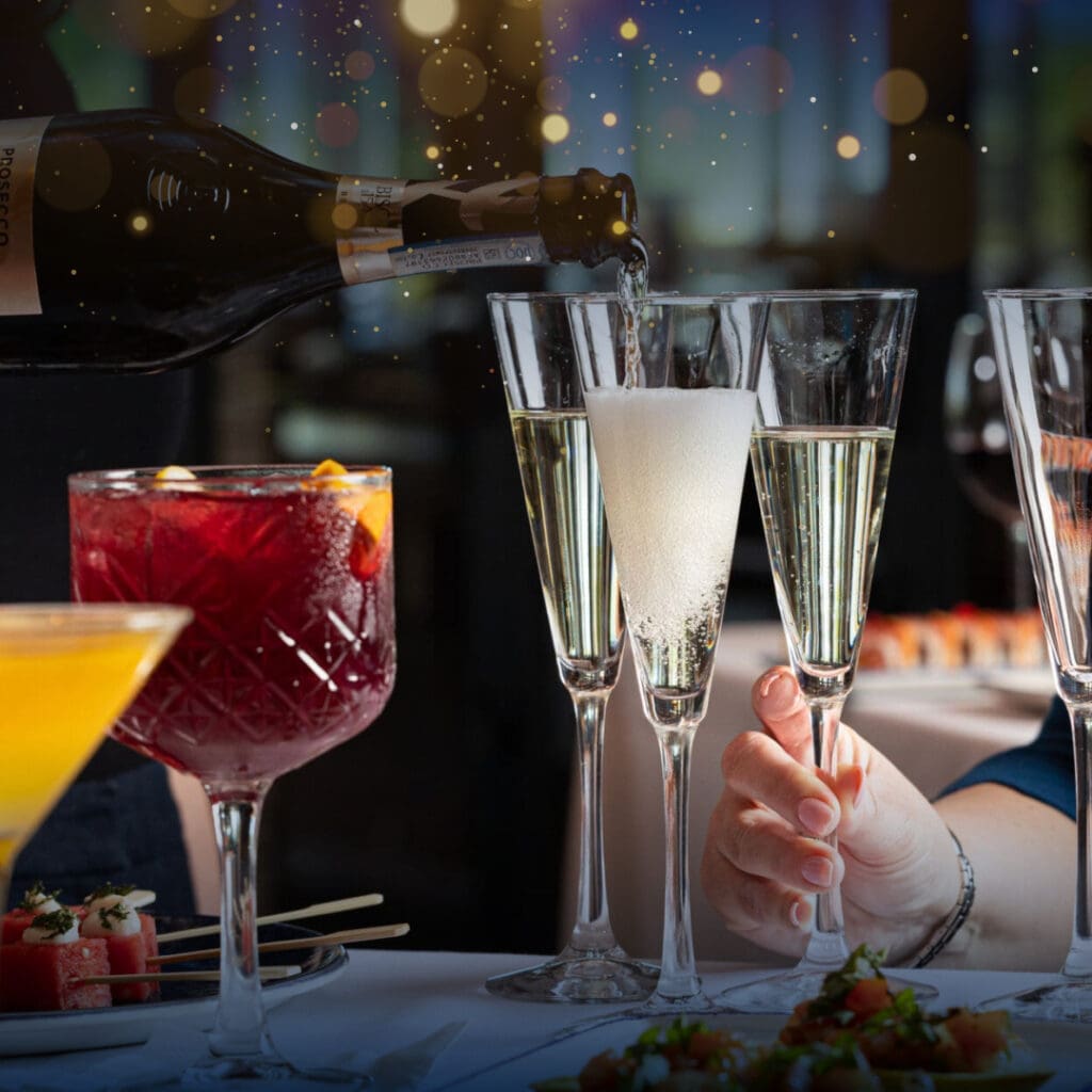 A hand holds a champagne flute as sparkling wine is poured. Nearby, glasses of red sangria and an orange cocktail are on the table. The setting has a festive atmosphere with blurred sparkling lights in the background.