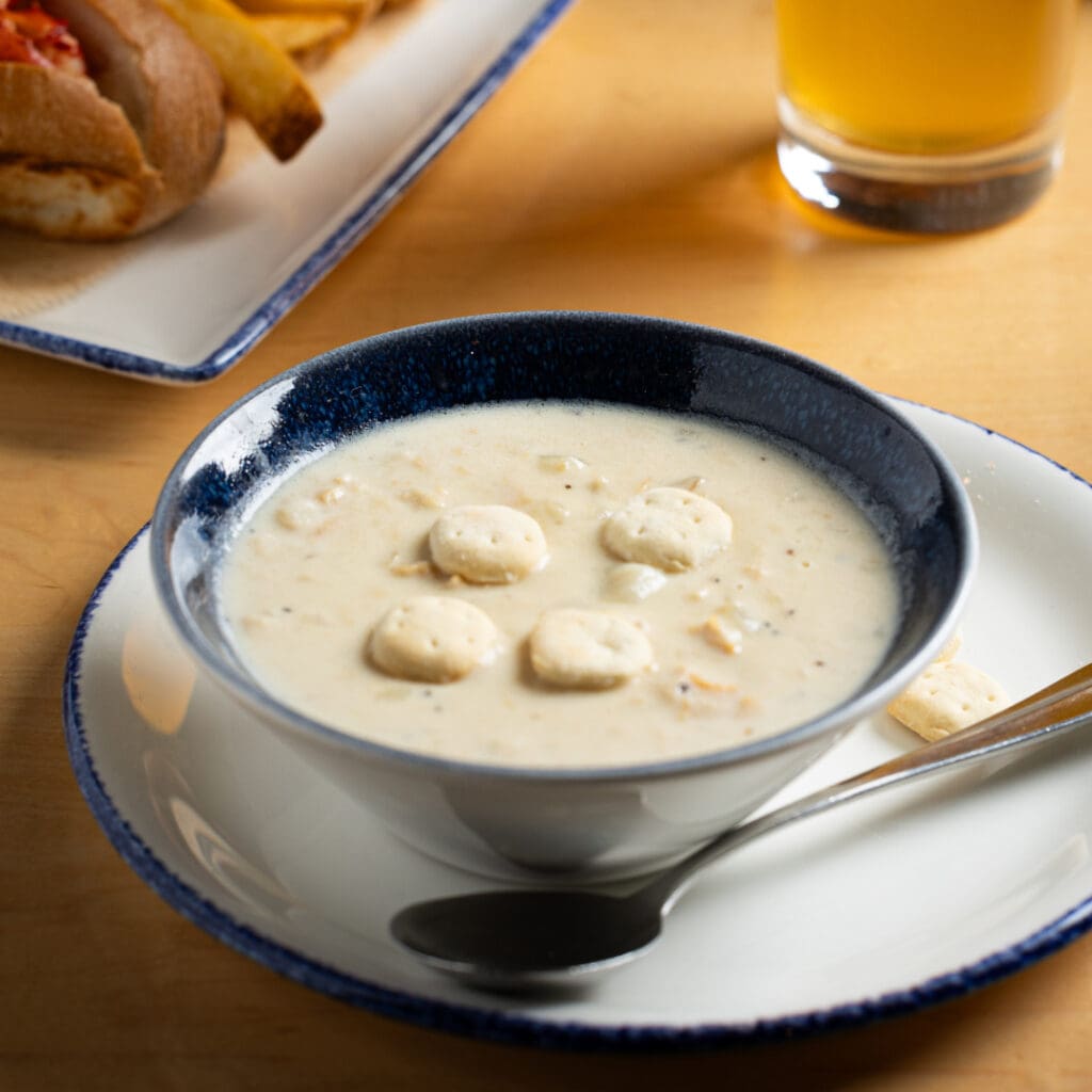 A bowl of creamy clam chowder topped with oyster crackers sits on a blue-rimmed plate with a spoon. In the background, a sandwich and a glass of orange-hued drink are visible. The setting is a wooden table.