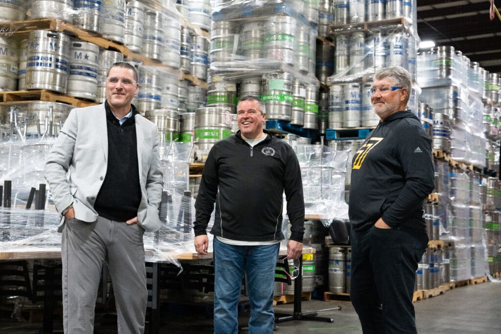 Three men are standing and smiling in a large warehouse filled with stacked metal kegs. They are dressed in casual to semi-formal clothing and appear to be engaged in conversation.