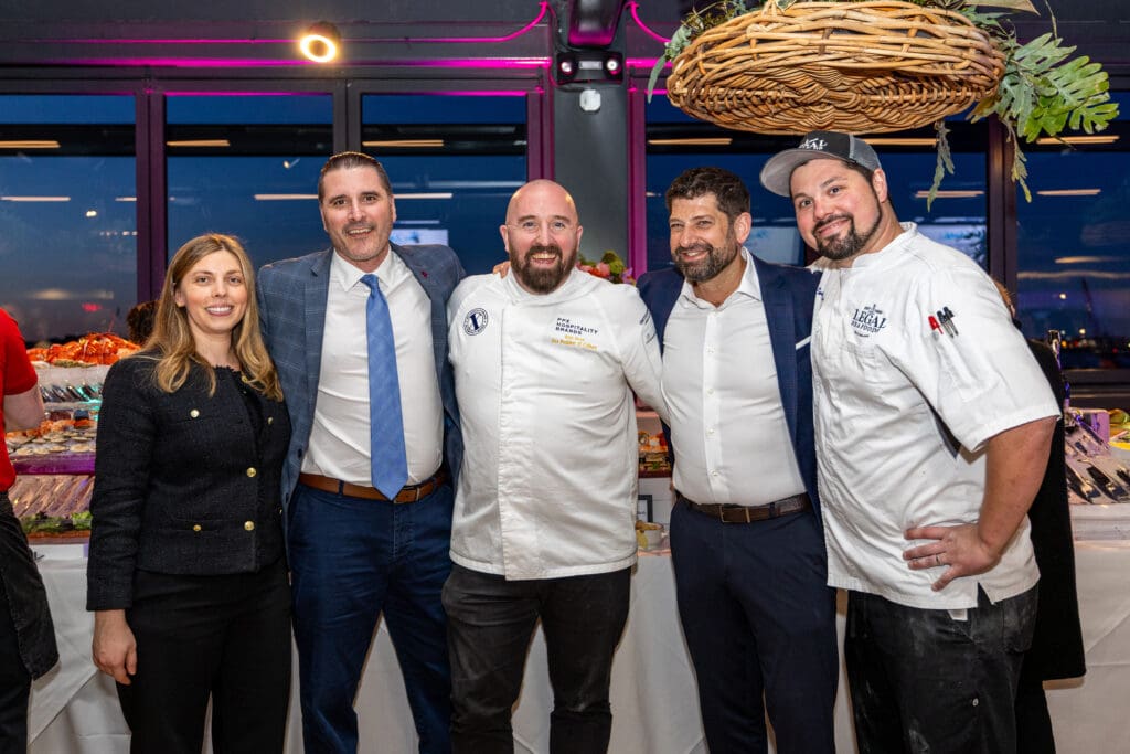 A group of five people, including four men and one woman, are smiling and posing together indoors. Two of the men are wearing chef uniforms, while the others are in business attire. A buffet table and city skyline are visible in the background.