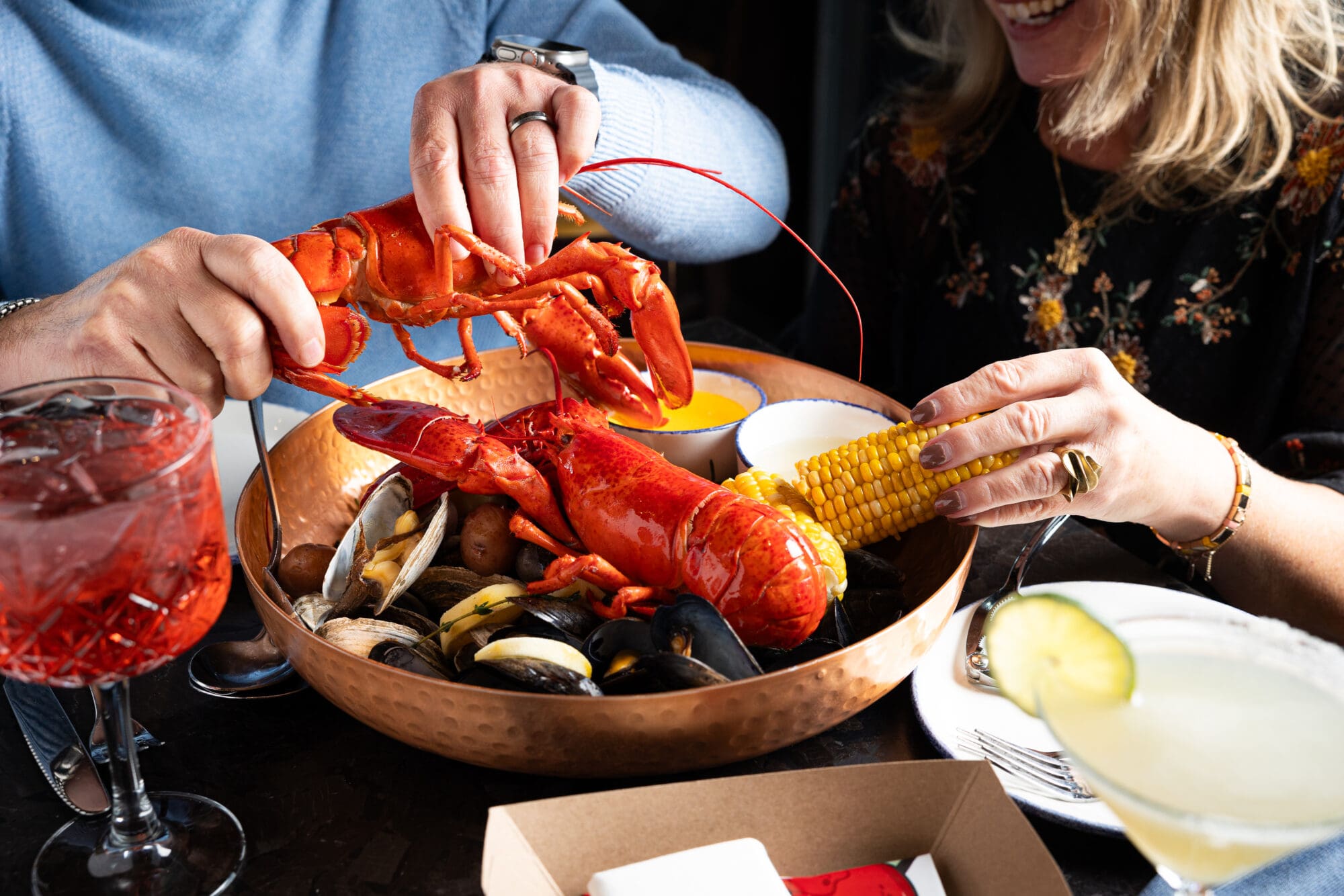 Two people enjoy a seafood feast, featuring whole lobsters, clams, and corn on the cob. Drinks, including a red cocktail and a margarita, are visible on the table, creating a lively dining atmosphere.