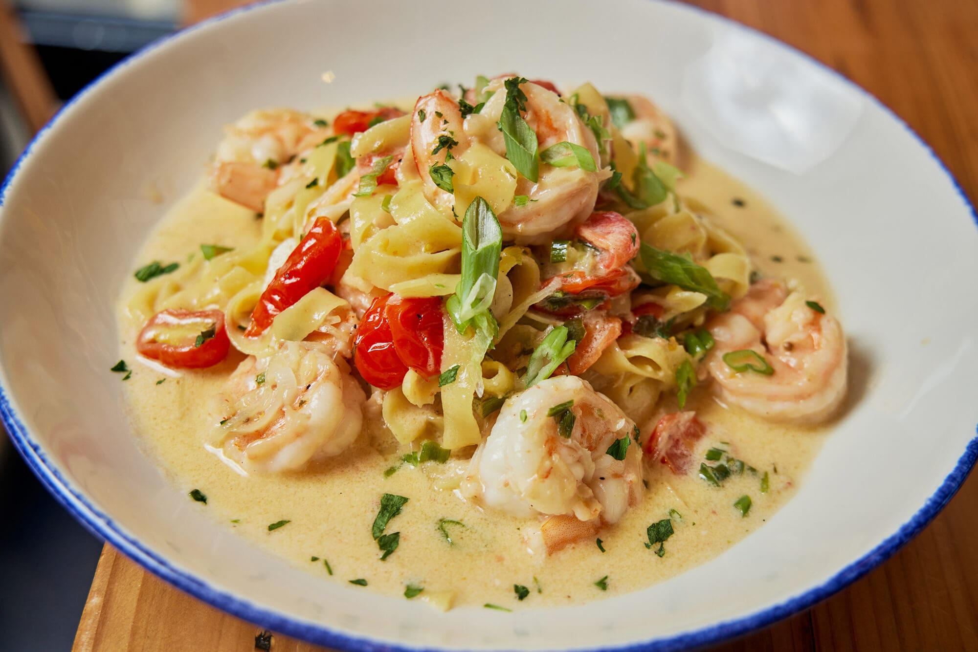 A creamy pasta dish featuring tagliatelle with shrimp, cherry tomatoes, and green onions, garnished with chopped herbs, is served in a white bowl with a blue rim.