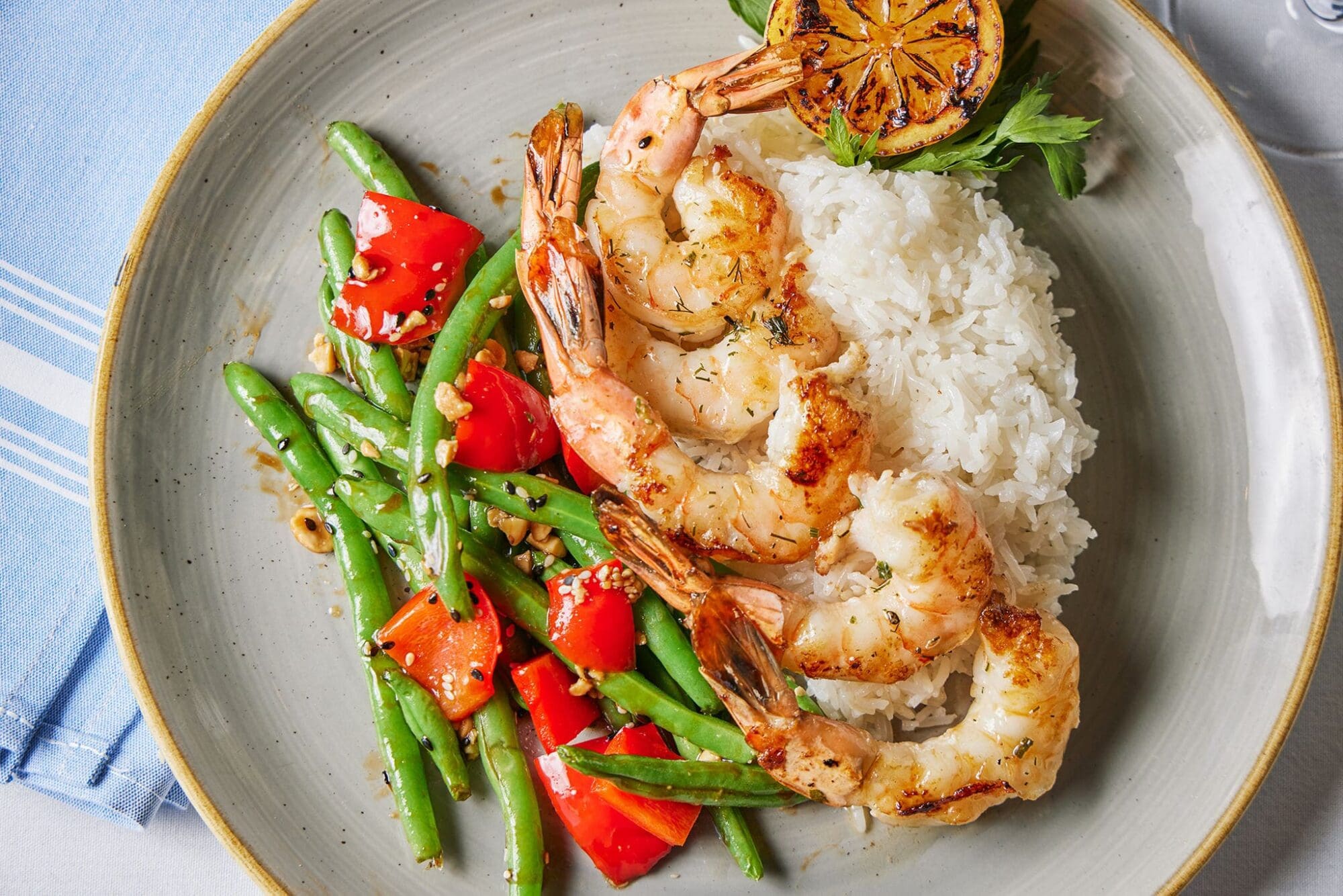 A plate of grilled shrimp, white rice, and a side of sautéed green beans and red bell peppers. A grilled lemon wedge is garnished on the side, all served on a light gray plate with a blue napkin underneath.