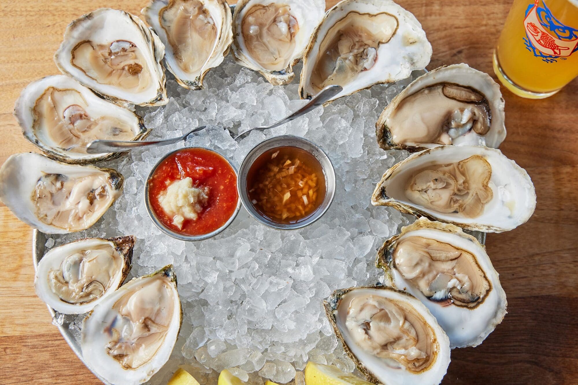 A plate of twelve opened oysters on ice, arranged in a circle with two metal cups of red sauce and mignonette in the center. Two lemon wedges are placed at the bottom, and a glass of orange beverage is in the top right corner.