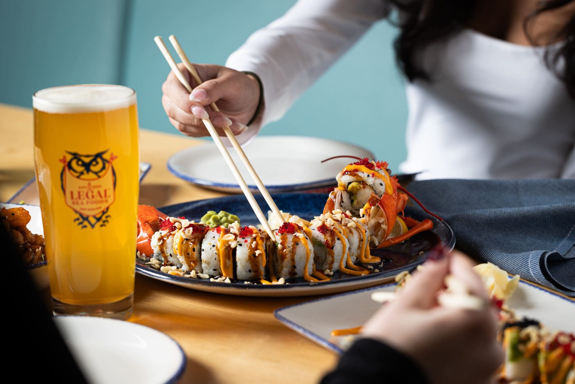 A person uses chopsticks to pick up sushi from a large plate on a wooden table. A tall glass of beer is next to the plate. Another person is partially visible in the foreground. Plates and napkins are also on the table.