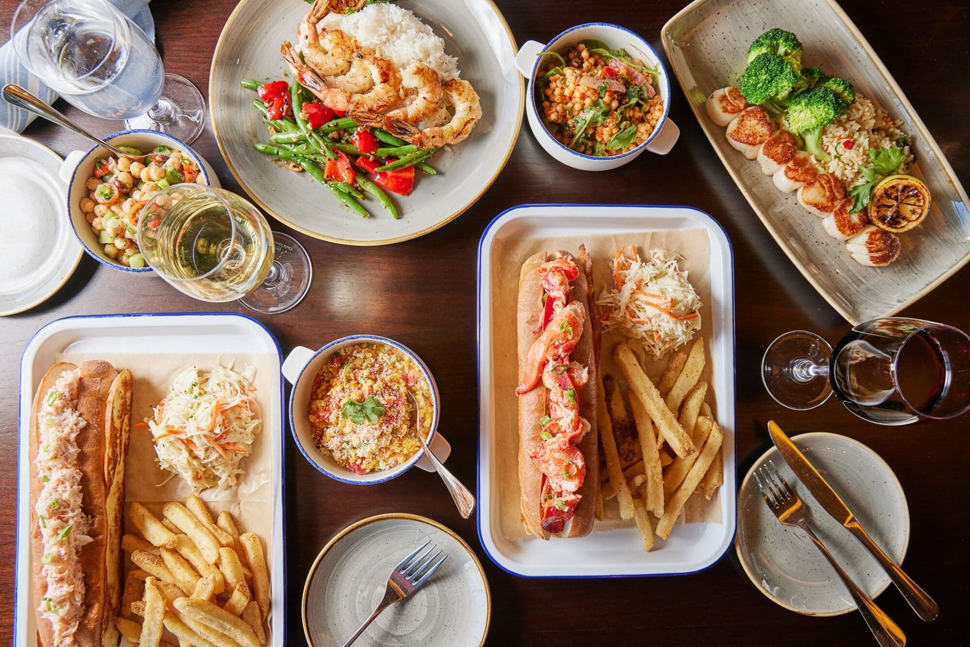 A table with a variety of dishes including lobster rolls with fries, seafood pasta, grilled vegetables, broccoli, quinoa salad, and two glasses of wine. Plates and utensils are arranged around the dishes.