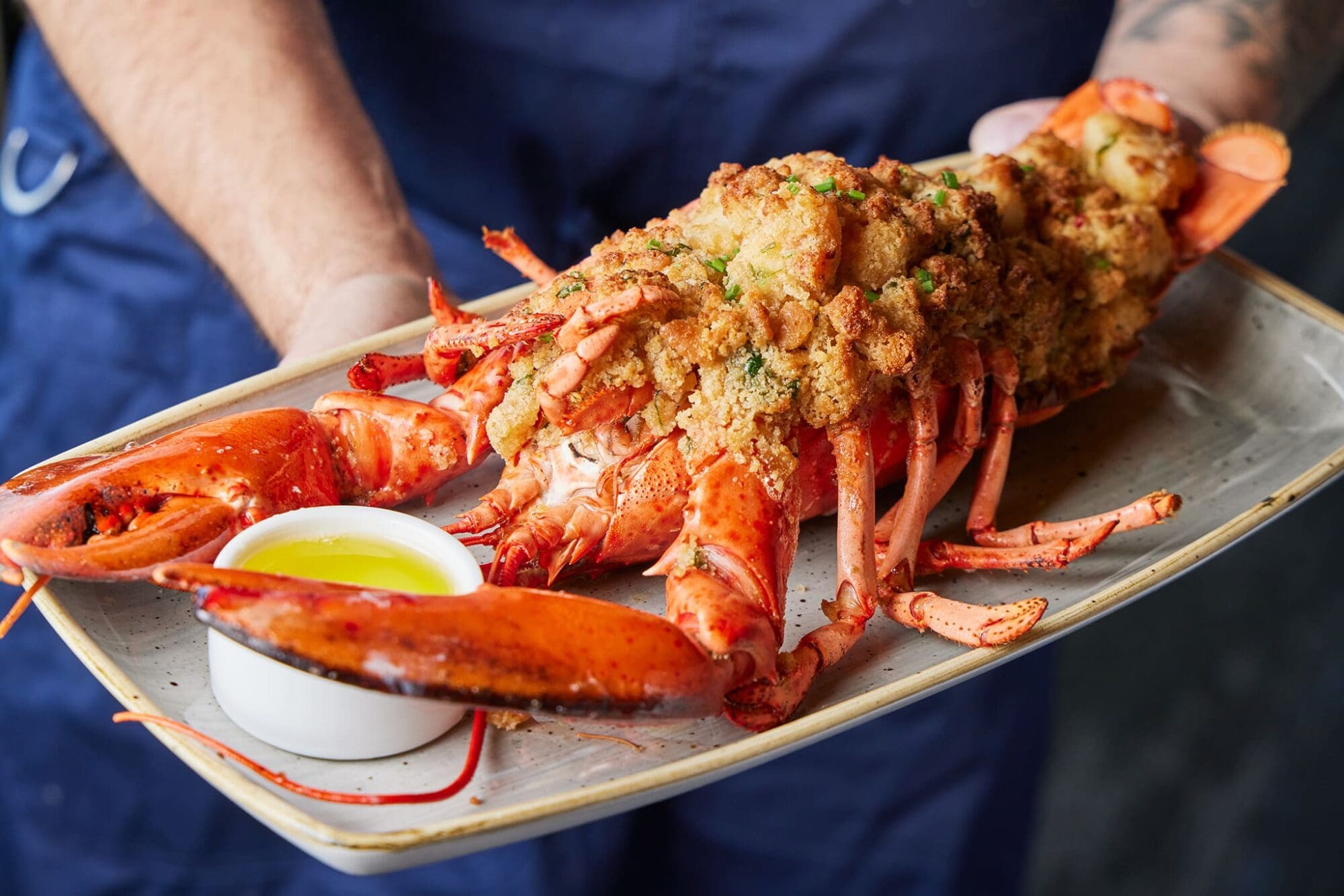 A person holding a platter with a stuffed lobster, served with a small bowl of melted butter. The lobster is garnished and appears freshly cooked, resting on a rectangular gray dish. The person's blue attire is partially visible in the background.