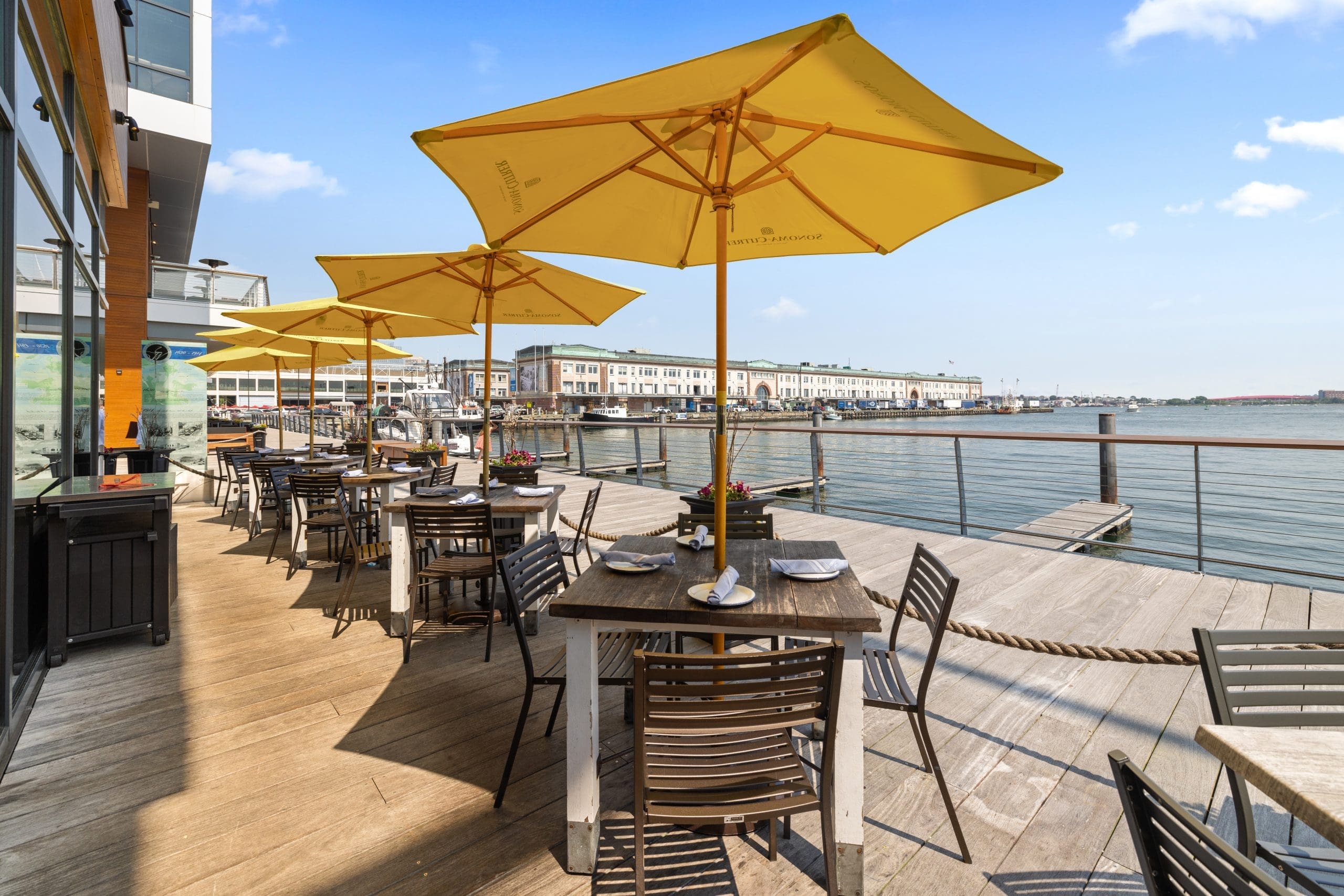 Outdoor dining area on a wooden deck by the water, featuring tables with black chairs and yellow umbrellas. A large building is visible in the background under a clear blue sky.