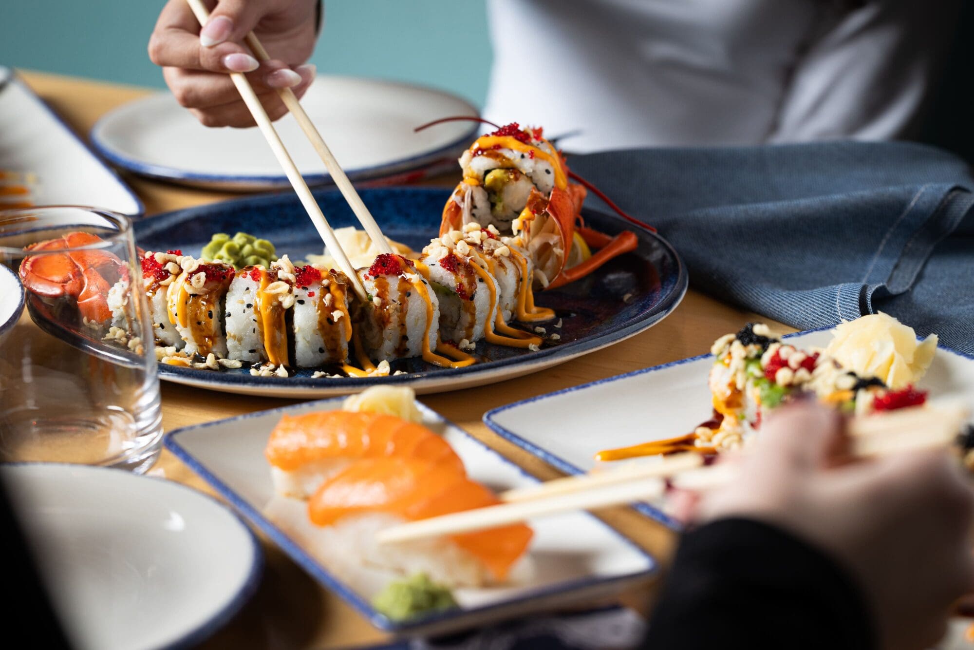 Close-up of people using chopsticks to pick up sushi from platters on a table. The plates include sushi rolls topped with sauce and roe, and slices of salmon. There's a glass of water and a folded blue napkin nearby.