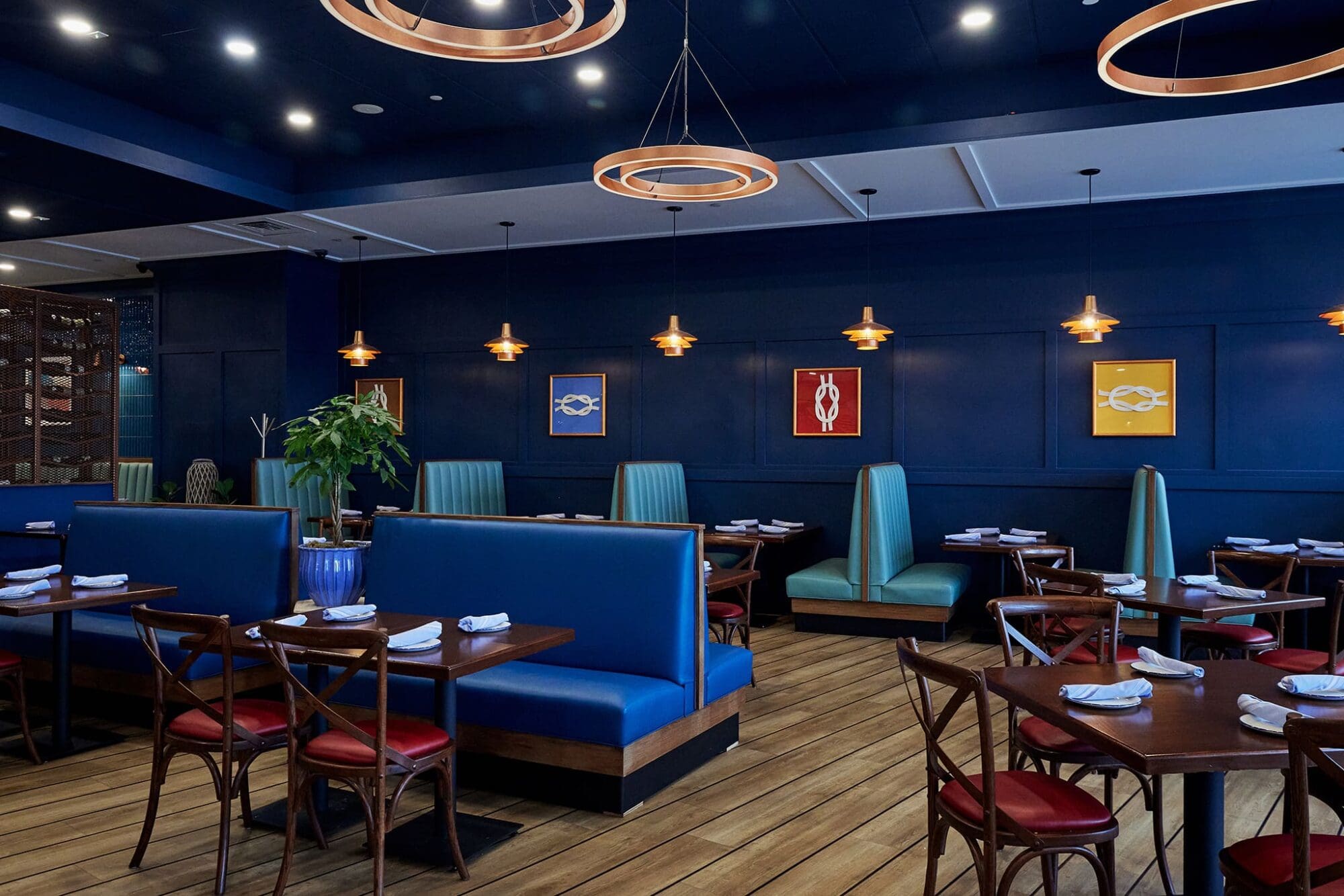 A chic restaurant interior with deep blue walls and wooden flooring. Modern pendant lights illuminate booths and wooden tables set with white napkins. Colorful fish-themed art decorates the walls. Green plants add a touch of nature.