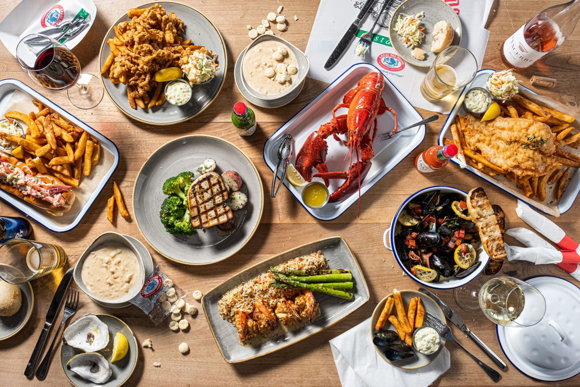 A wooden table is filled with various seafood dishes, including a whole lobster, grilled fish, fried shrimp, oysters, and mussels. Side dishes like fries, coleslaw, and lemon slices accompany the seafood, with glasses of white wine nearby.