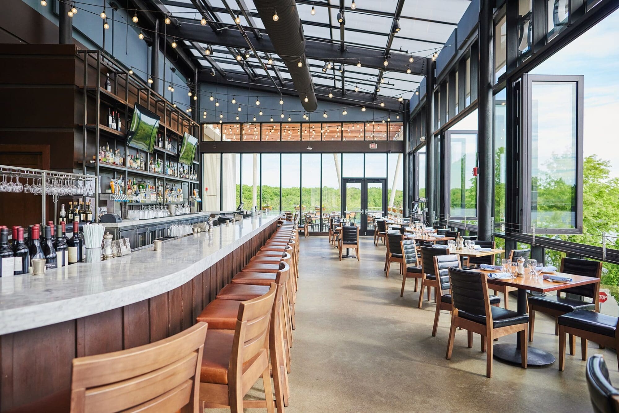 Spacious restaurant interior with a long marble bar lined with brown chairs, shelves stocked with bottles, and tables with black chairs. Large windows provide a view of greenery outside. Ceiling features string lights and a modern design.