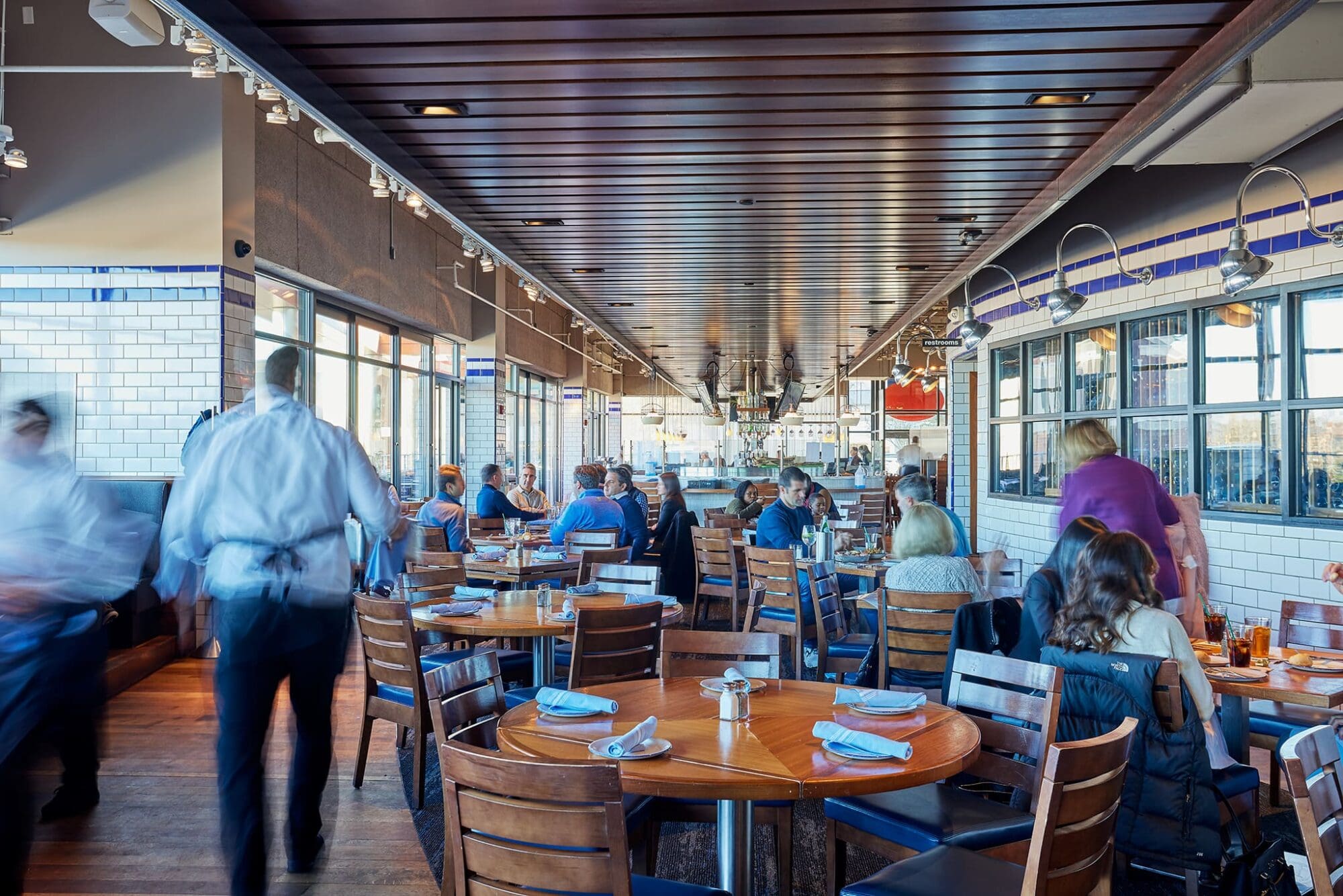 A bustling, modern restaurant with wooden tables and chairs. Diners are seated, eating, and socializing. Waitstaff in motion blur attend to guests. The room is bright with natural light from large windows.