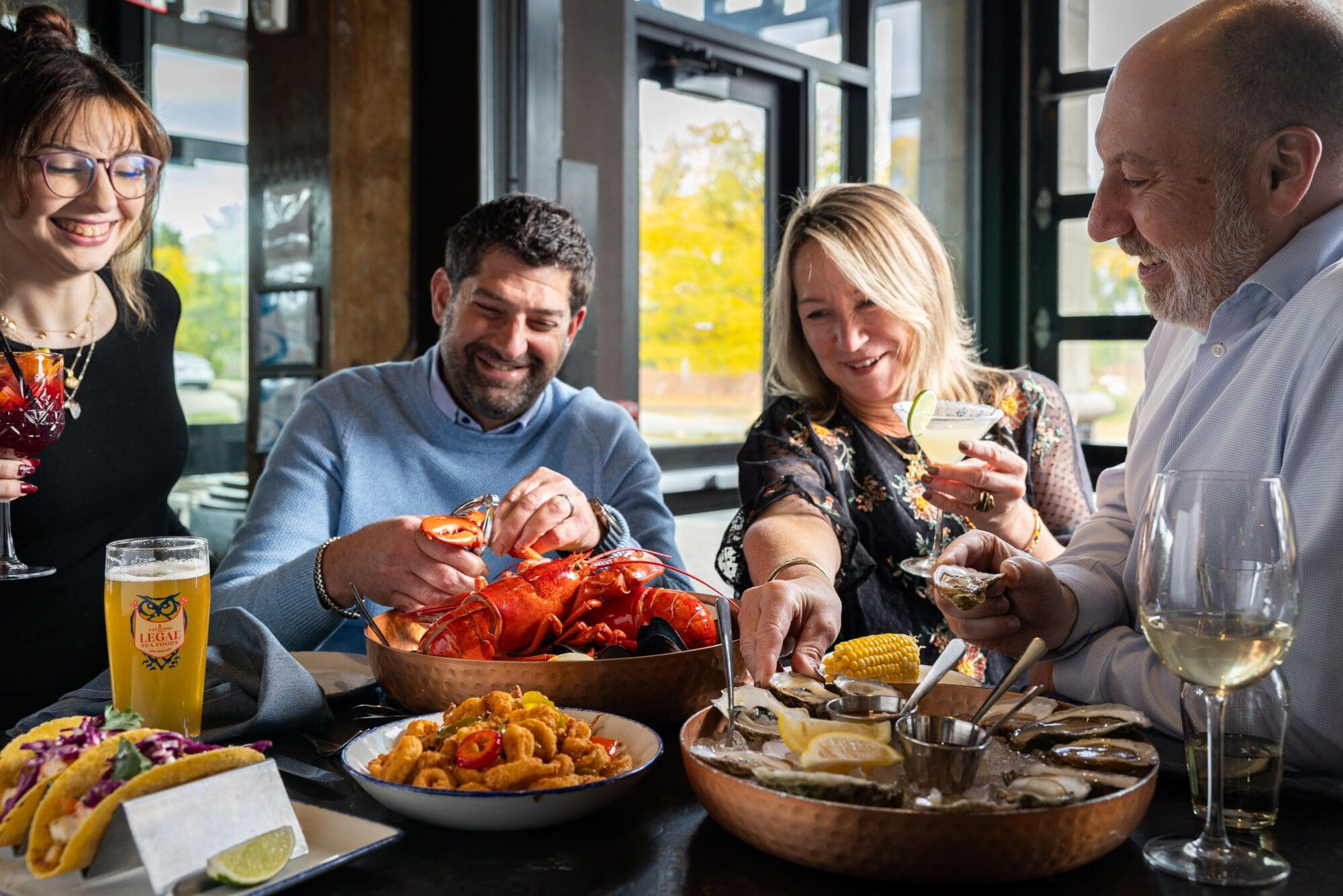 Four people enjoy a seafood meal at a restaurant, featuring lobsters, oysters, and drinks. They are laughing and interacting, creating a convivial atmosphere. The table is set near a window with natural light.