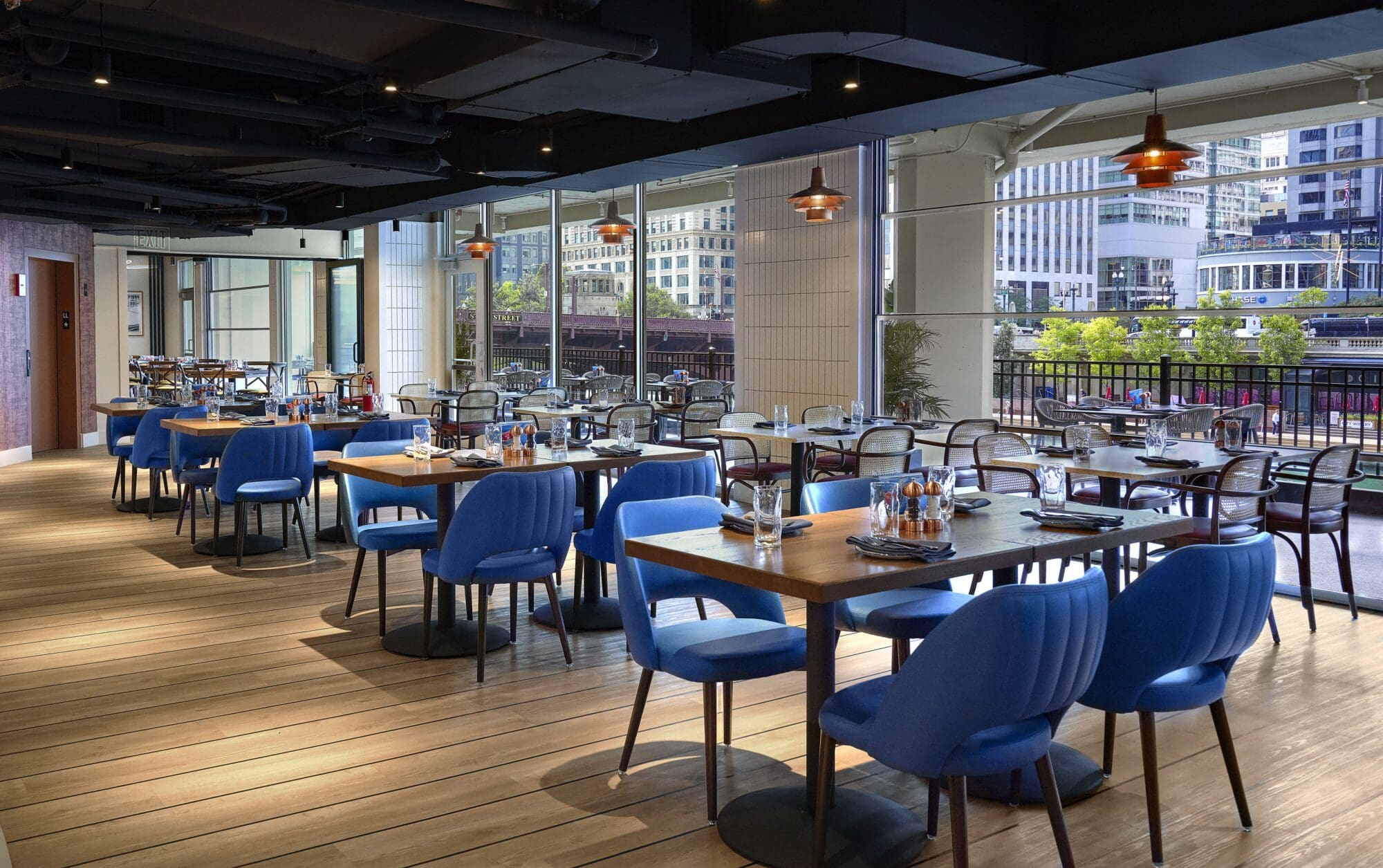 Spacious modern restaurant interior with wooden flooring, featuring blue chairs and wooden tables set with glasses and cutlery. Large windows reveal a view of a cityscape with tall buildings and greenery outside.