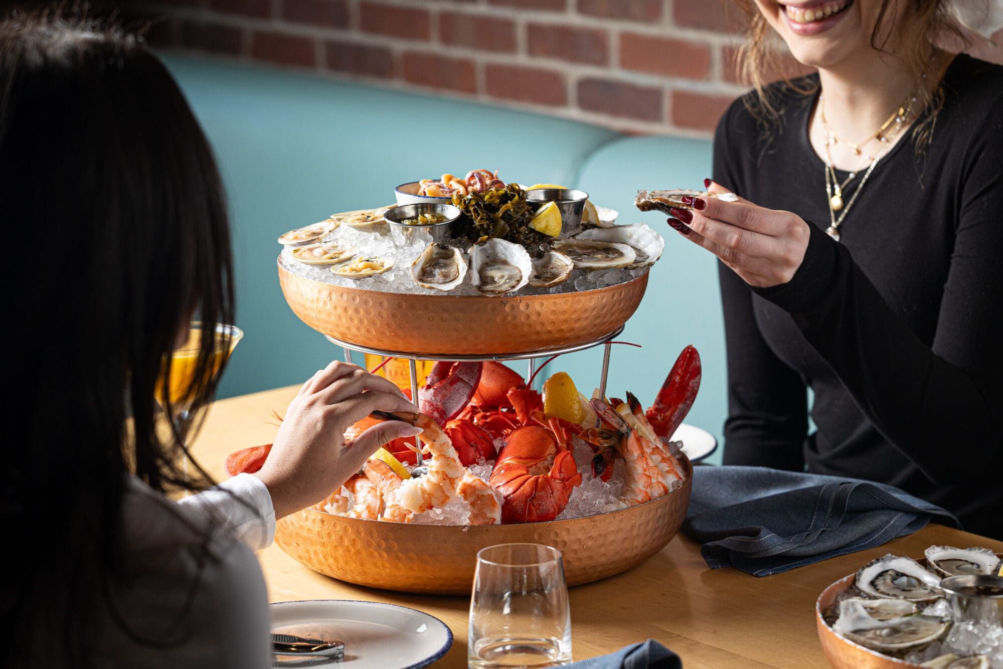 Two people enjoying a seafood tower in a restaurant. The tower includes oysters, lobster, and shrimp on ice, garnished with lemon wedges. The setting is casual with a brick wall background, teal seating, and glasses on the table.