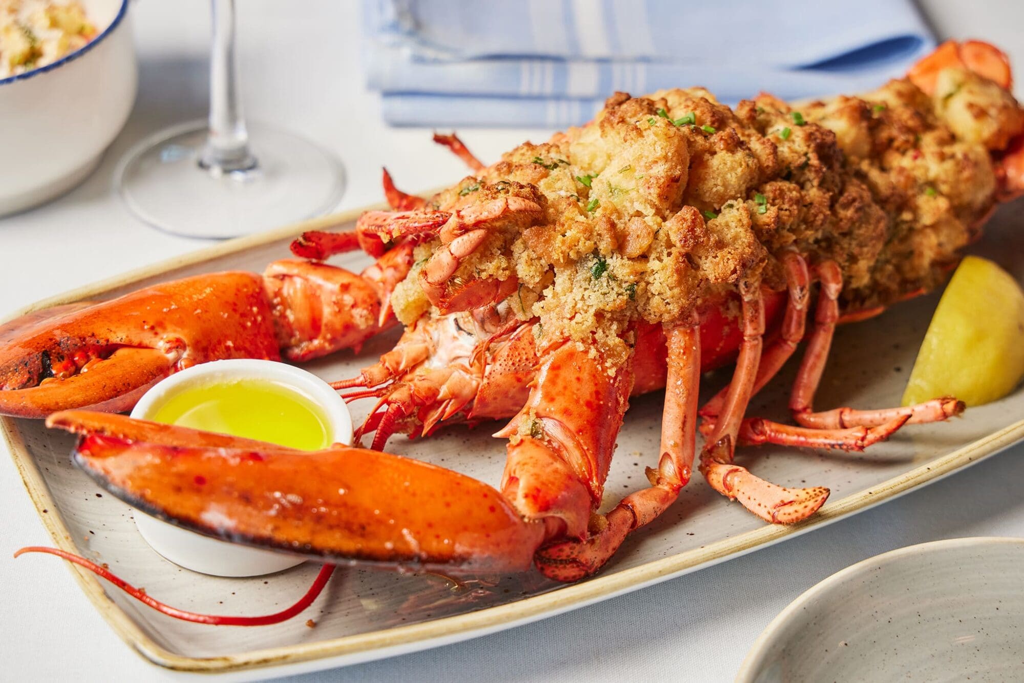 A stuffed lobster topped with a breadcrumb mixture is on a rectangular plate. A wedge of lemon and a small cup of melted butter are beside it. A glass of white wine and a blue-striped napkin are in the background.