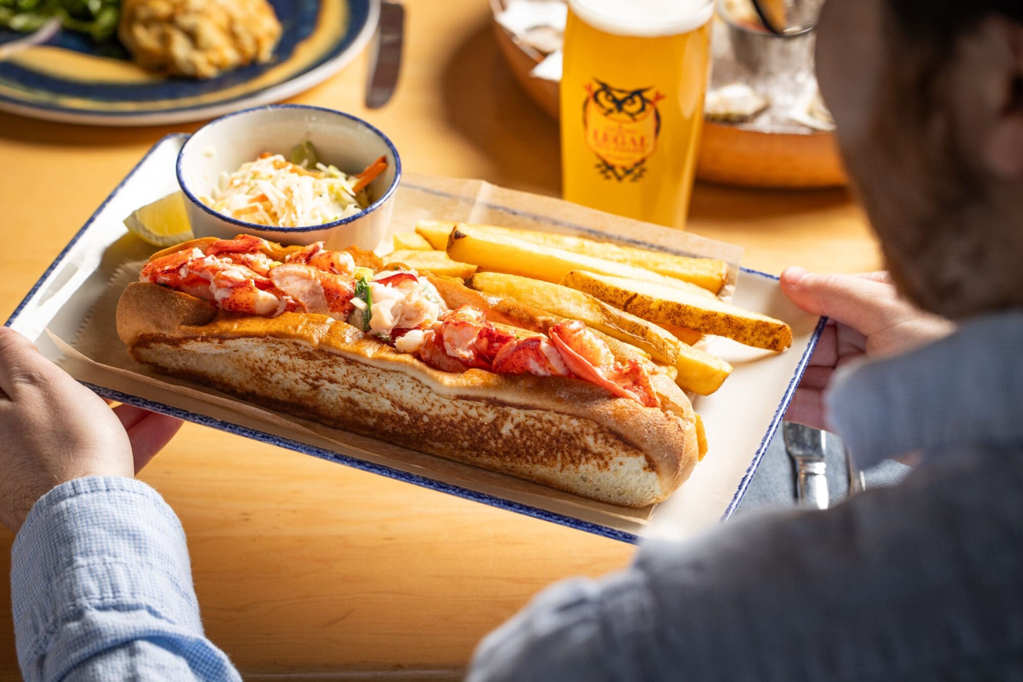 A person holding a plate with a lobster roll, fries, and a small bowl of coleslaw. A glass with a logo is in the background on the table. The setting appears to be a restaurant with wooden tables.